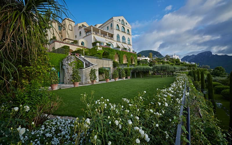 Elegant white villa with terraced gardens, manicured lawns, and white flowers against a mountain backdrop.