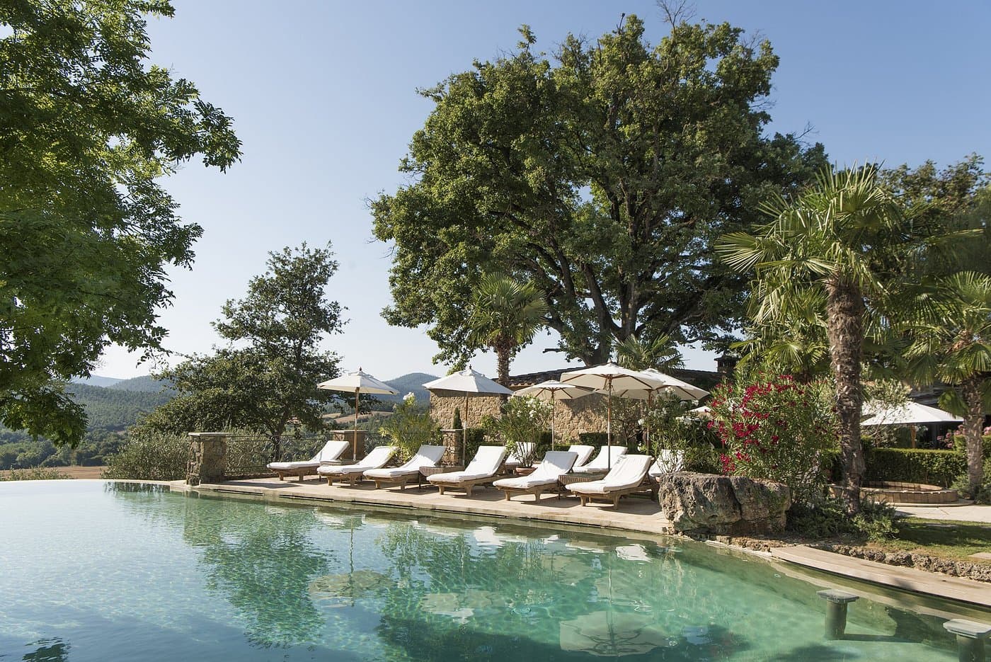 Infinity pool with white lounge chairs and umbrellas surrounded by lush trees and hills.