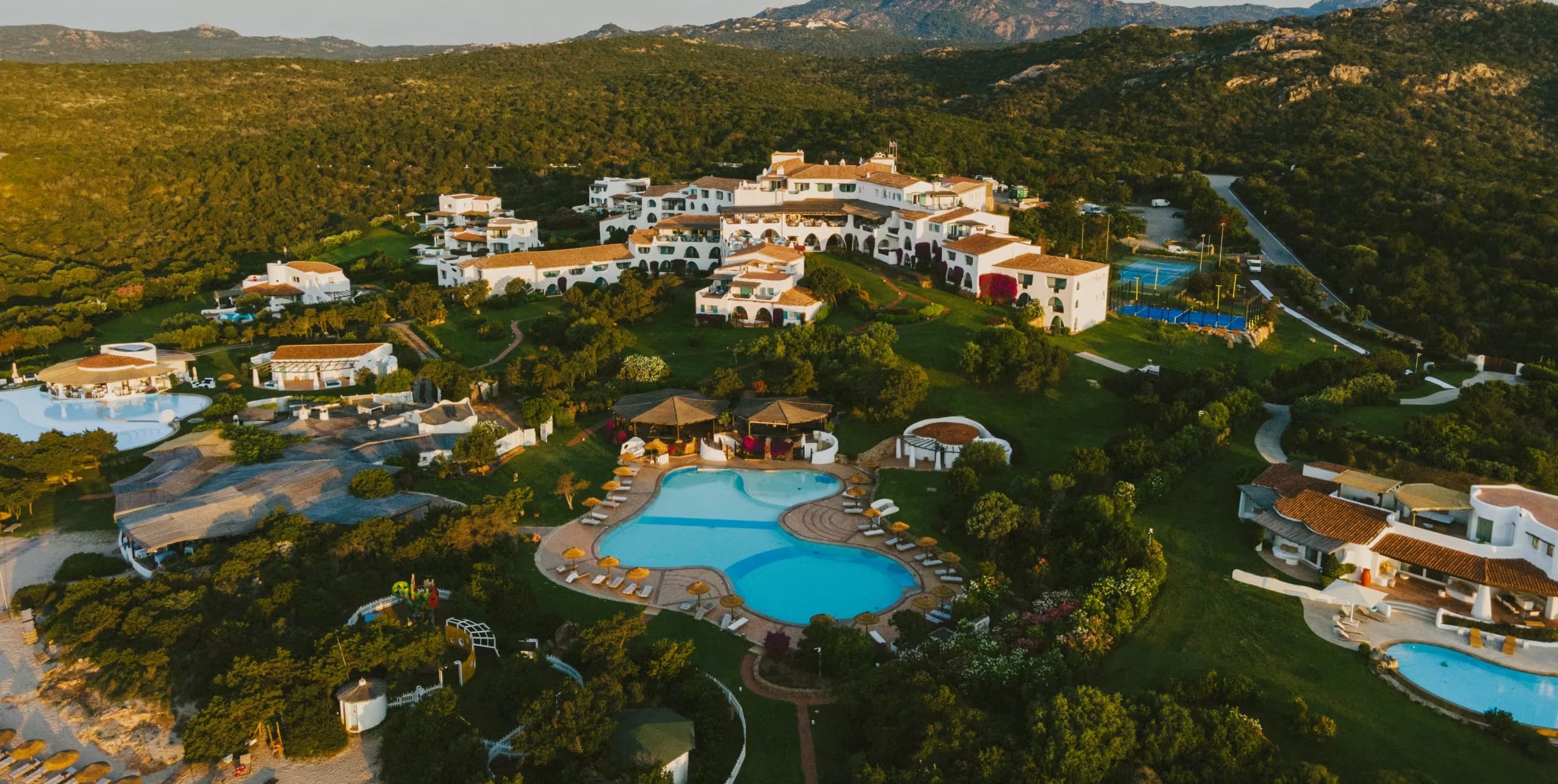 Sprawling luxury resort with white buildings and blue pools nestled in lush green hills.