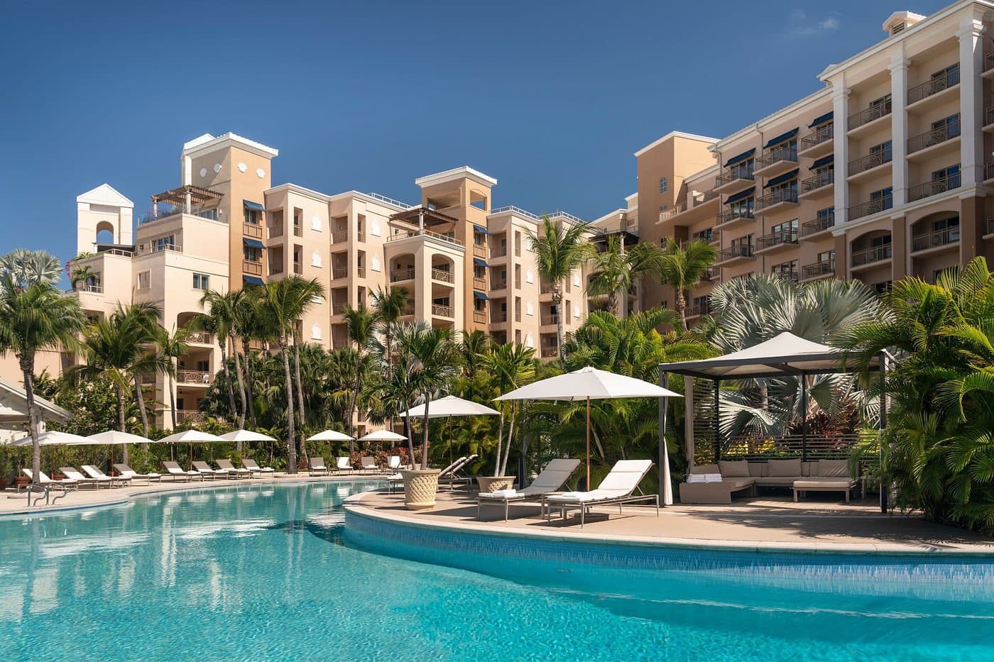 Luxury resort building overlooking a large swimming pool with umbrellas, lounge chairs, and palm trees.