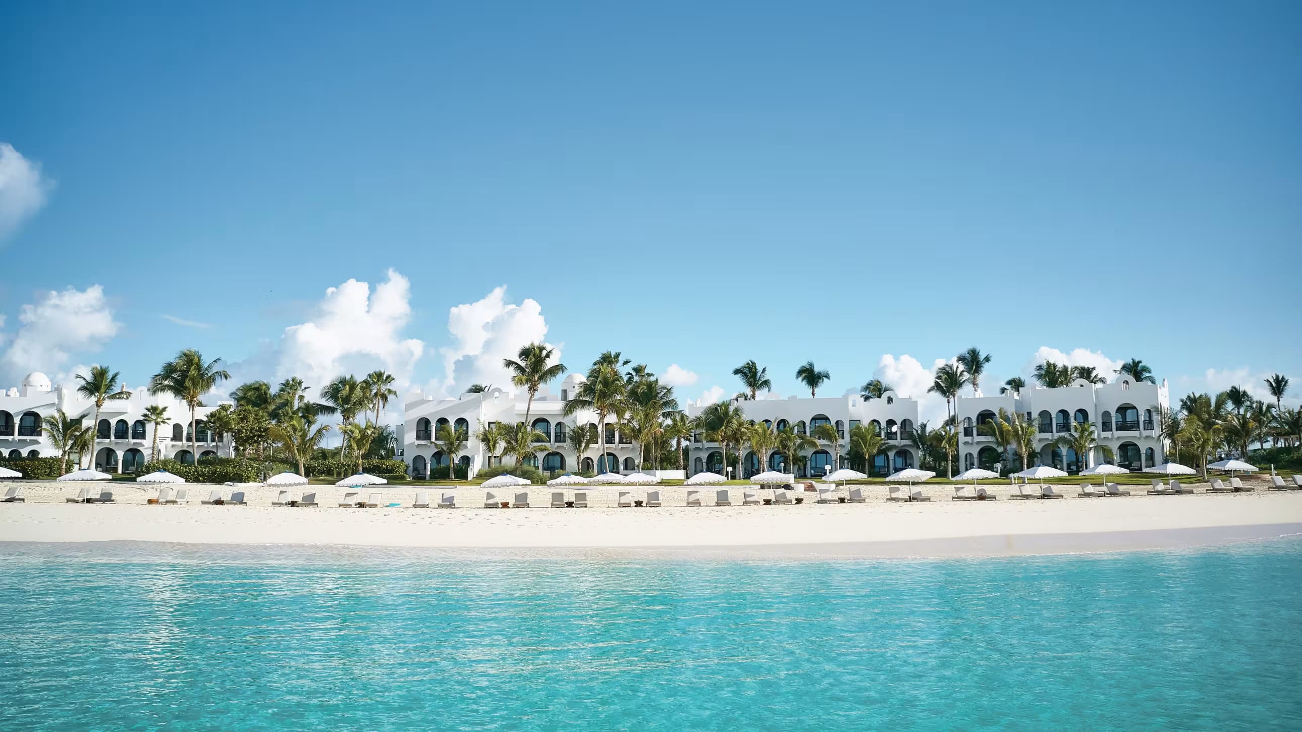 White Mediterranean-style resort buildings on a tropical beach with palm trees and turquoise water.