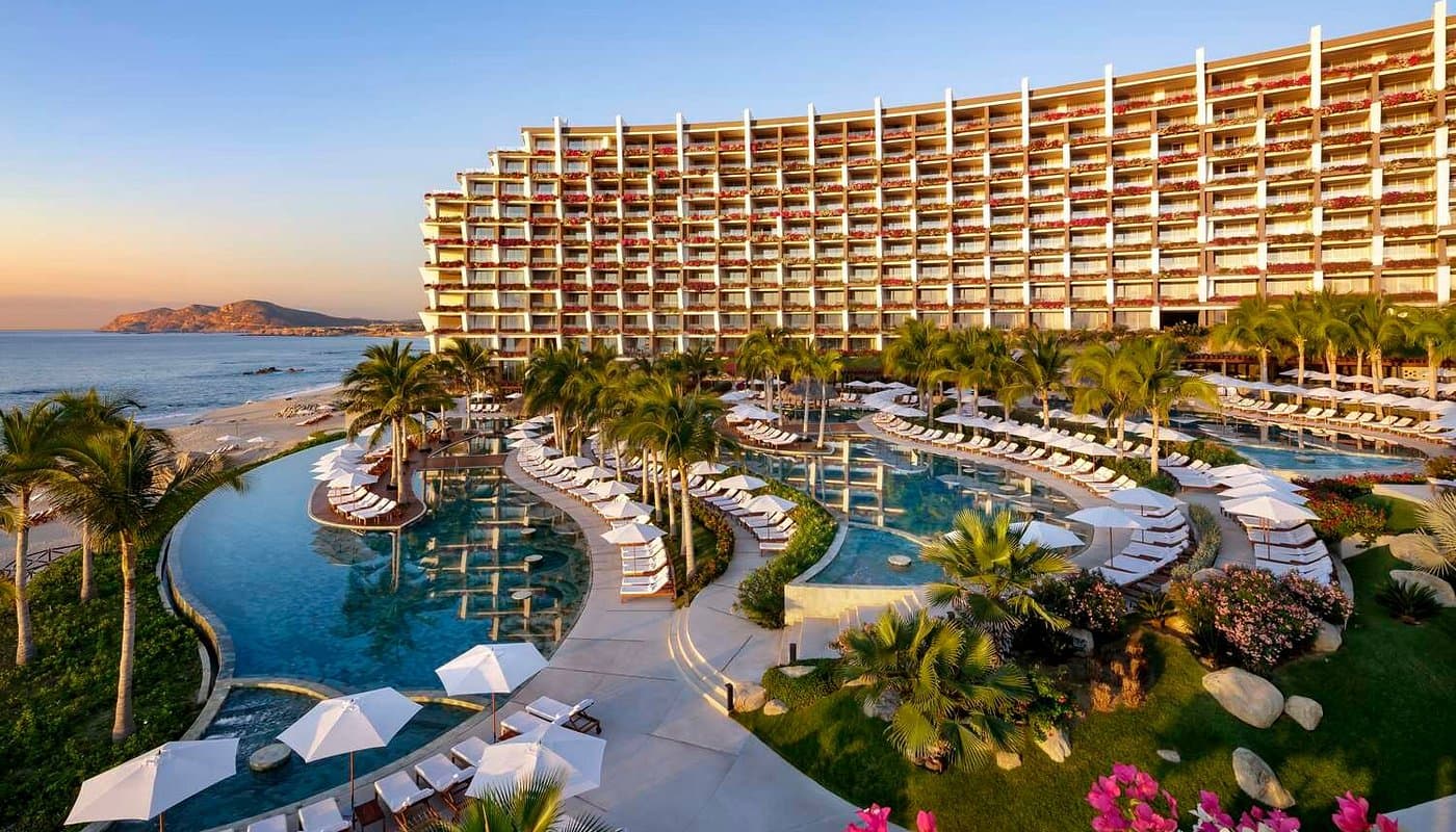 Luxury beachfront resort featuring tiered swimming pools, palm trees, and white umbrellas during sunset.