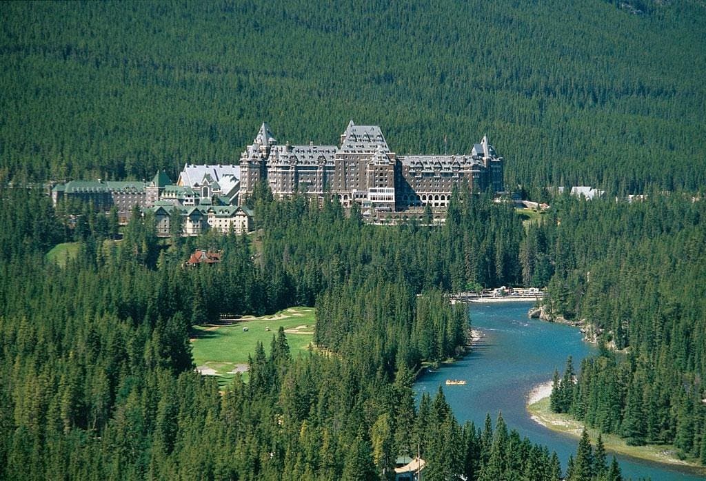 Grand stone hotel nestled in a dense evergreen forest beside a winding blue river.