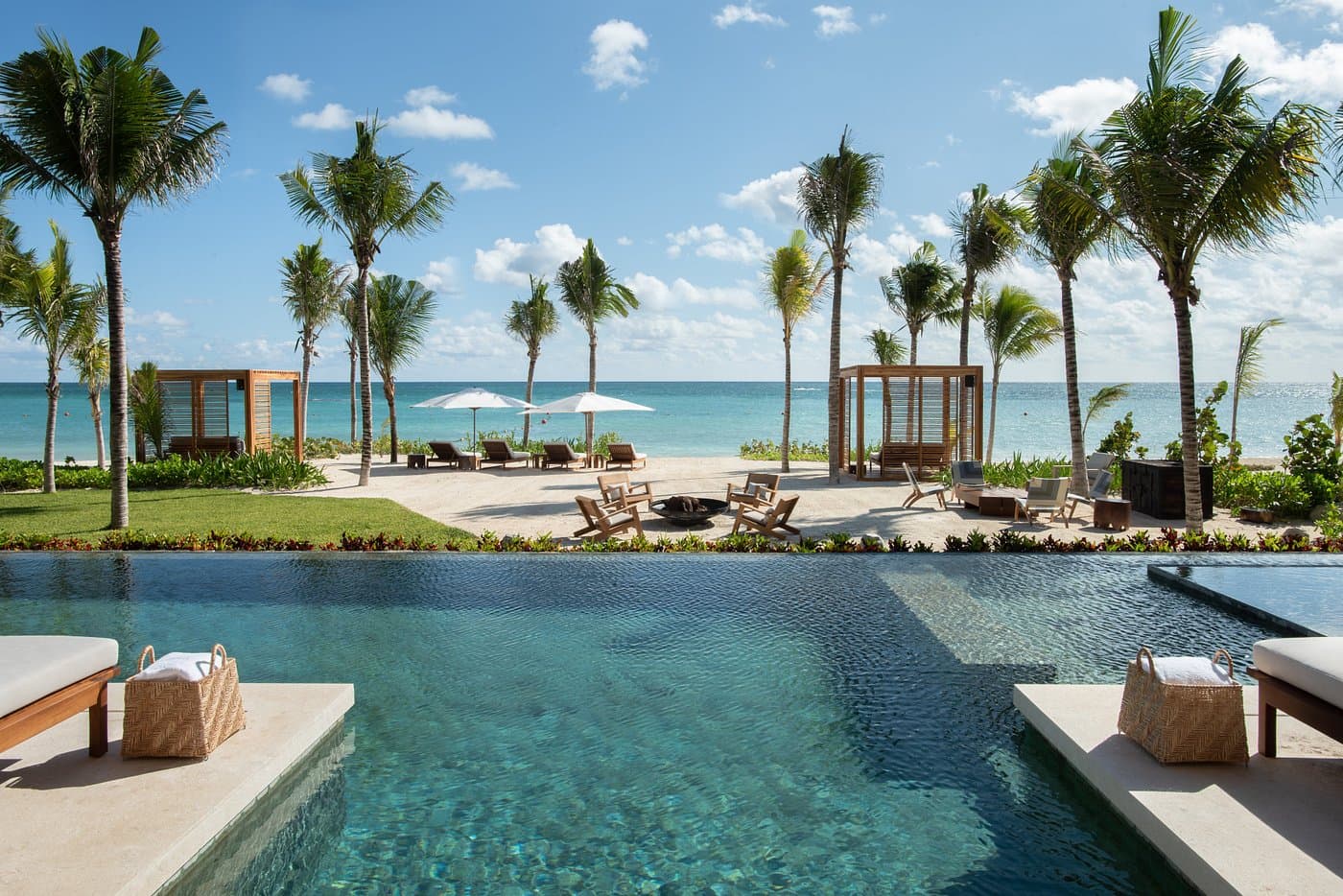 Luxury swimming pool overlooking a tropical beach with palm trees, cabanas, and turquoise ocean water.